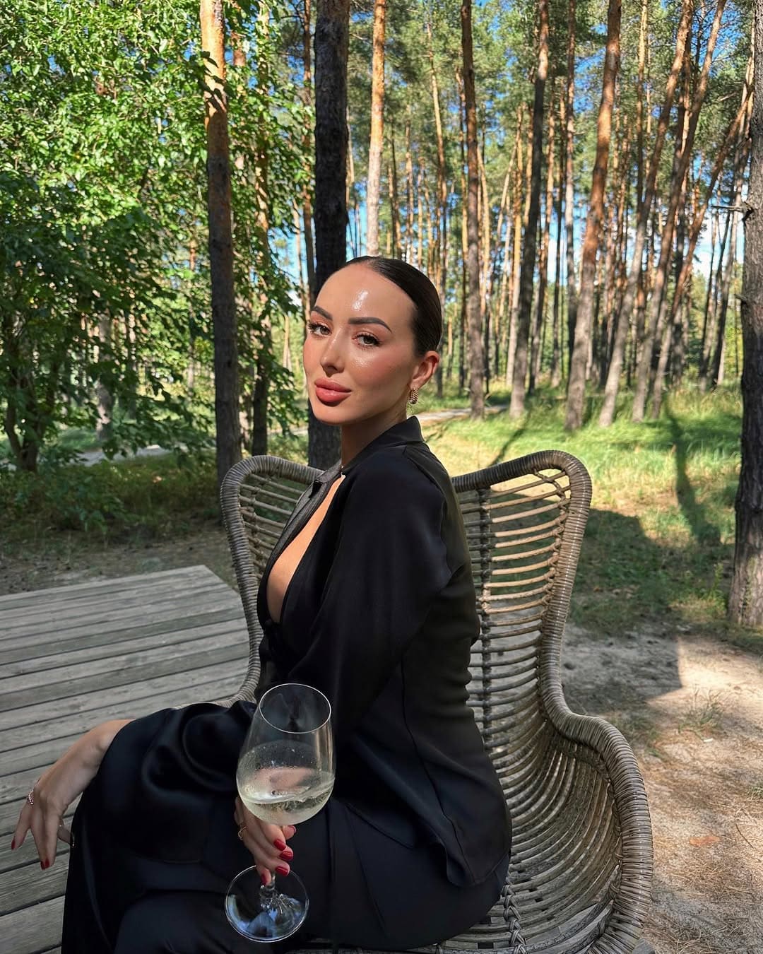 Mujer de vestido negro de manga larga sentada en silla de mimbre en bosque soleado.
