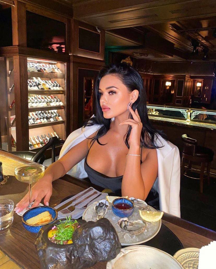 Renata con cabello oscuro largo sentada en mesa de restaurante con ostras y cóctel, usando top oscuro y chaqueta blanca.