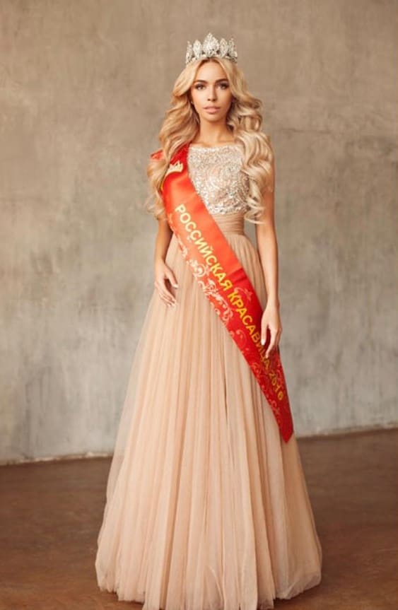 Natali TOP de frente con tiara, vestido champán con falda de tul y banda roja dorada, ante una pared texturizada.