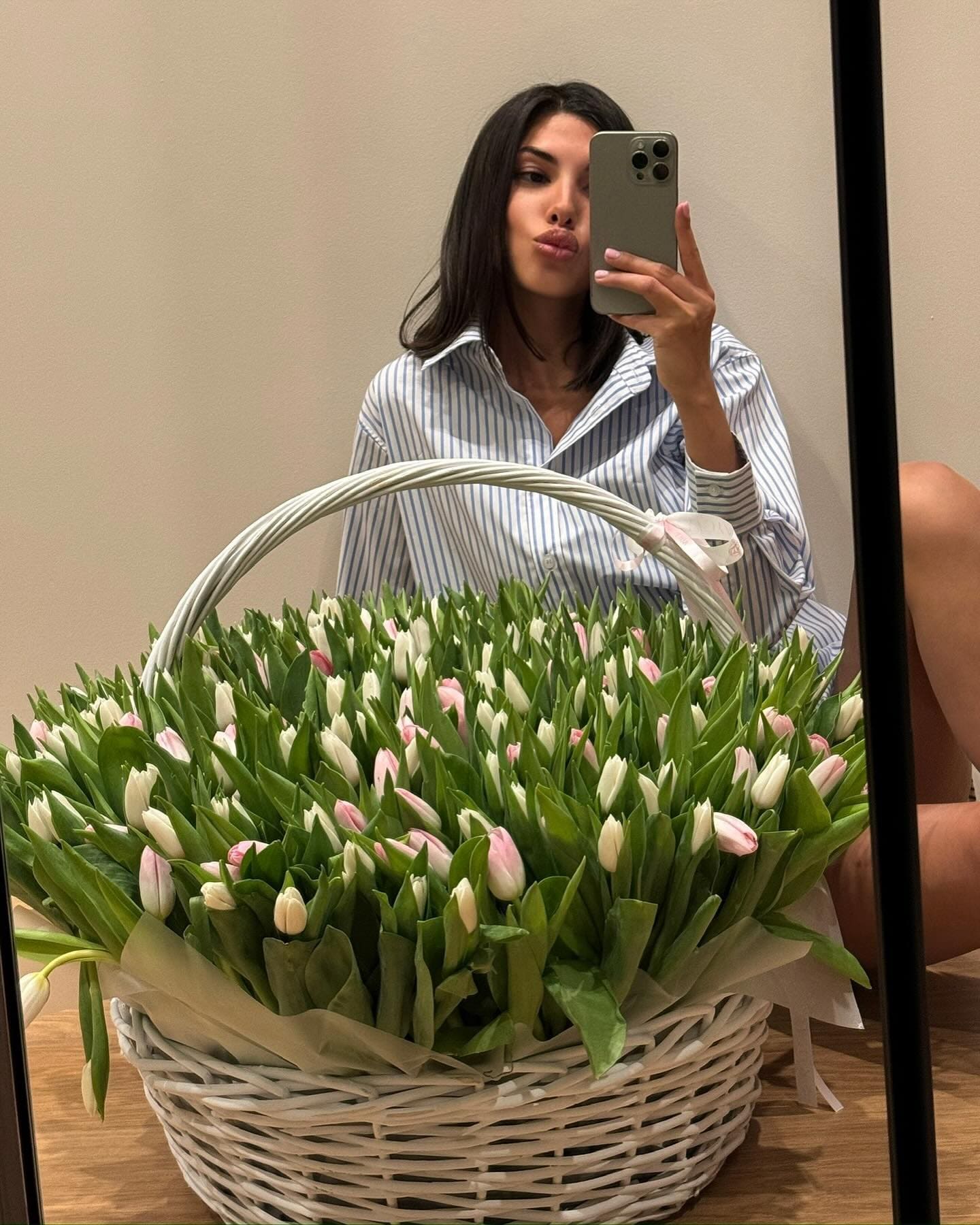 Mujer con cabello largo oscuro sentada en el suelo de madera frente a un espejo, vistiendo una camisa azul y blanca rayada grande, haciendo una selfie con los labios fruncidos.