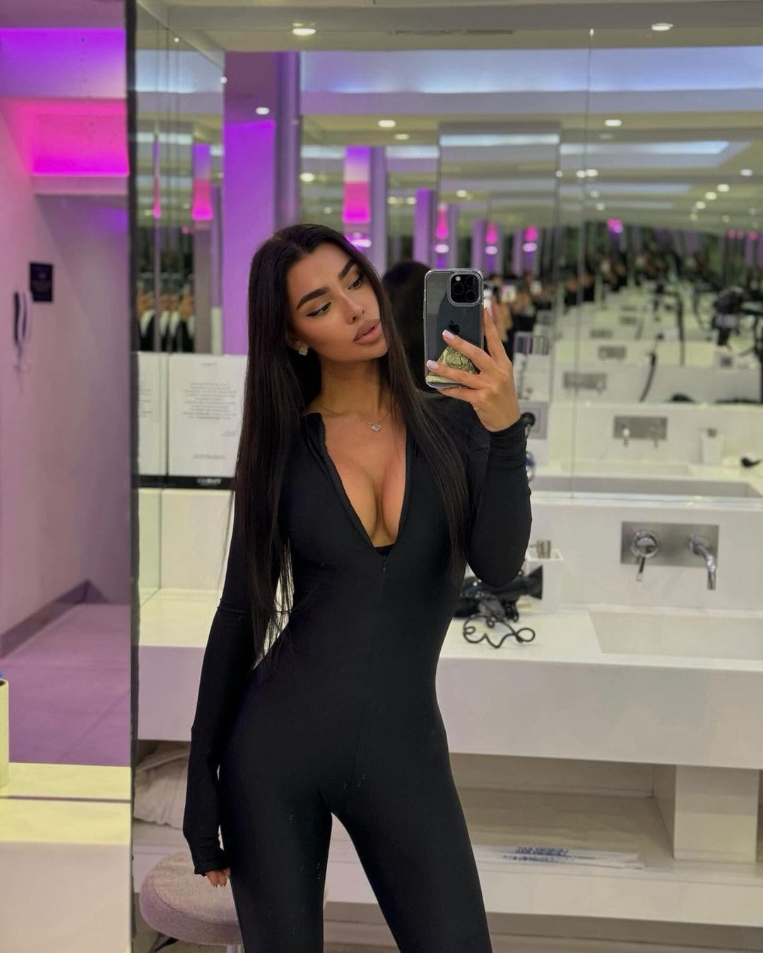 Mujer con cabello oscuro toma selfie en espejo con bodysuit negro de manga larga y escote V profundo, en habitación iluminada con luz púrpura.