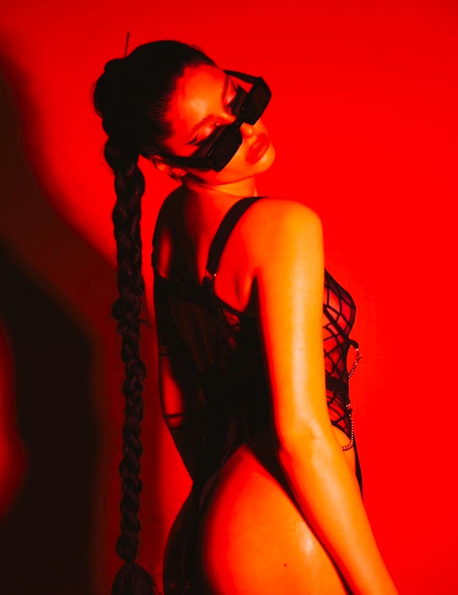 Perfil de una mujer con trenza larga y gafas grandes, vistiendo un bodysuit negro ajustado sobre fondo rojo.