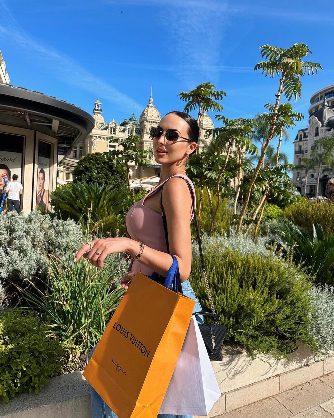 Mujer de perfil con top rosa y vaqueros sosteniendo bolsas de compra en jardín urbano.