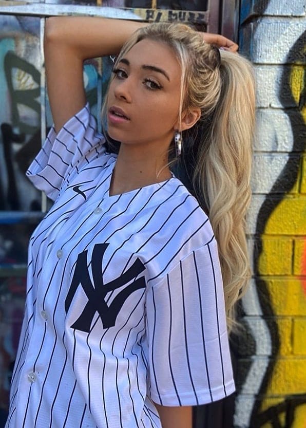 Rubia con coleta alta y manos tras la cabeza, viste camisa blanca rayada con logo NY ante ladrillo pintado.