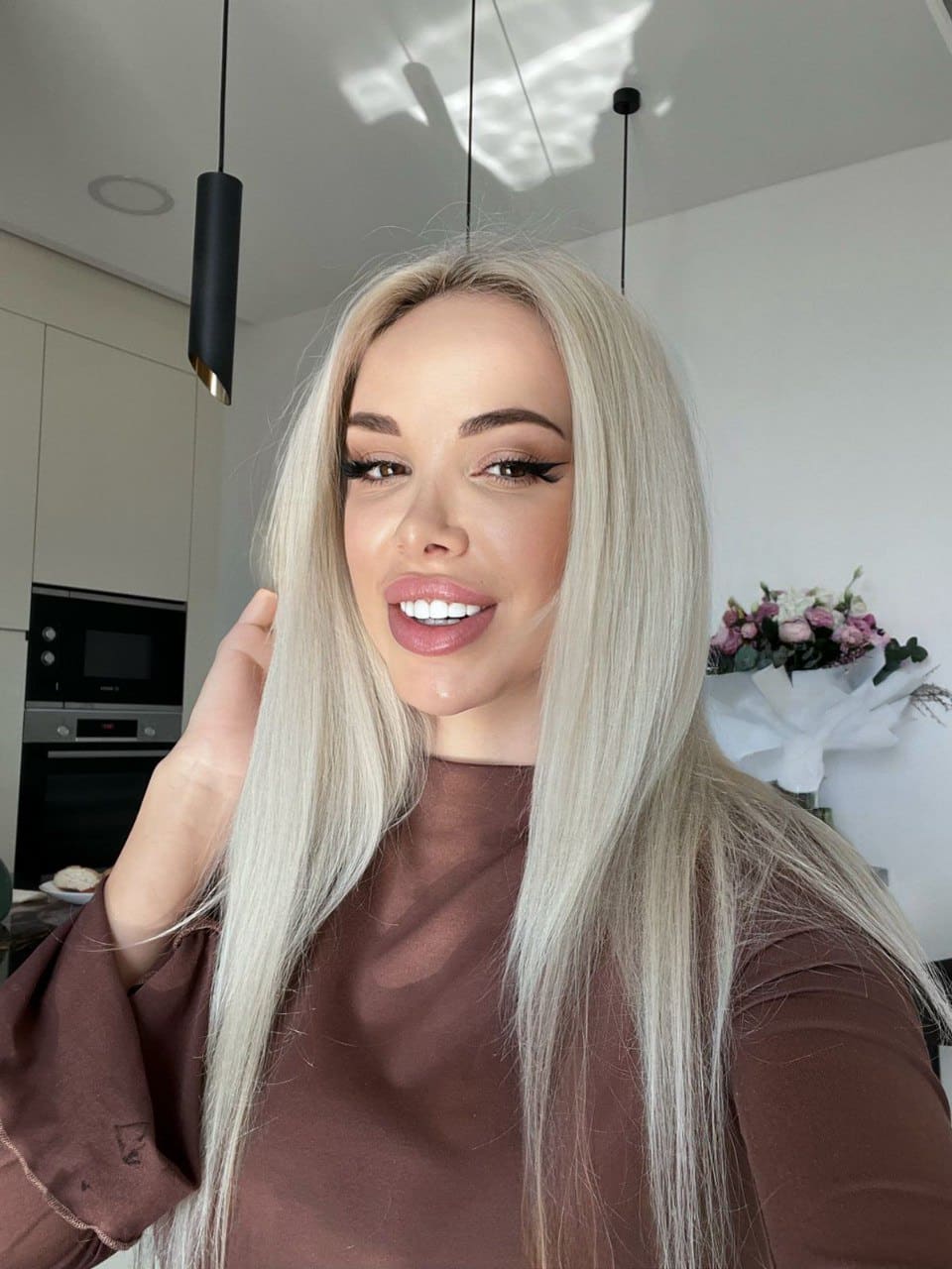 Platinum-blonde hair frames a brown long-sleeved top as one hand is raised near the face in a kitchen with white cabinets.