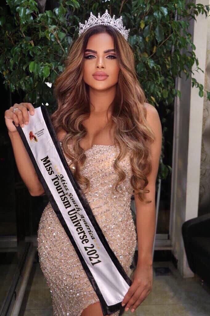 Sasha con cabello largo rizado castaño de pie al aire libre frente a follaje verde, vestida con un vestido sin tirantes brillante y una diadema.