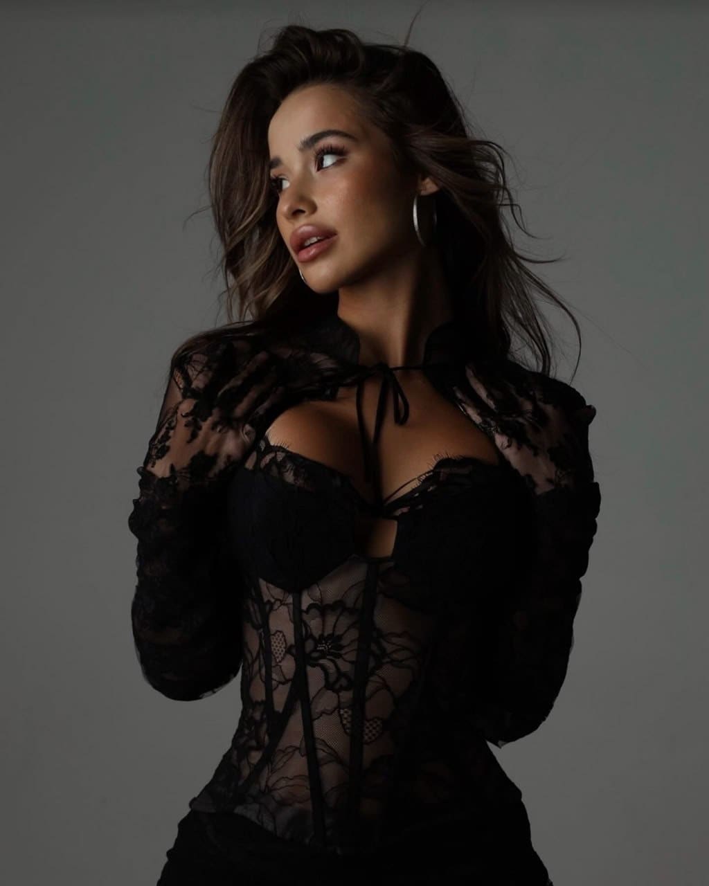 A woman stands in a studio wearing a black lace long-sleeved top with an open neckline.