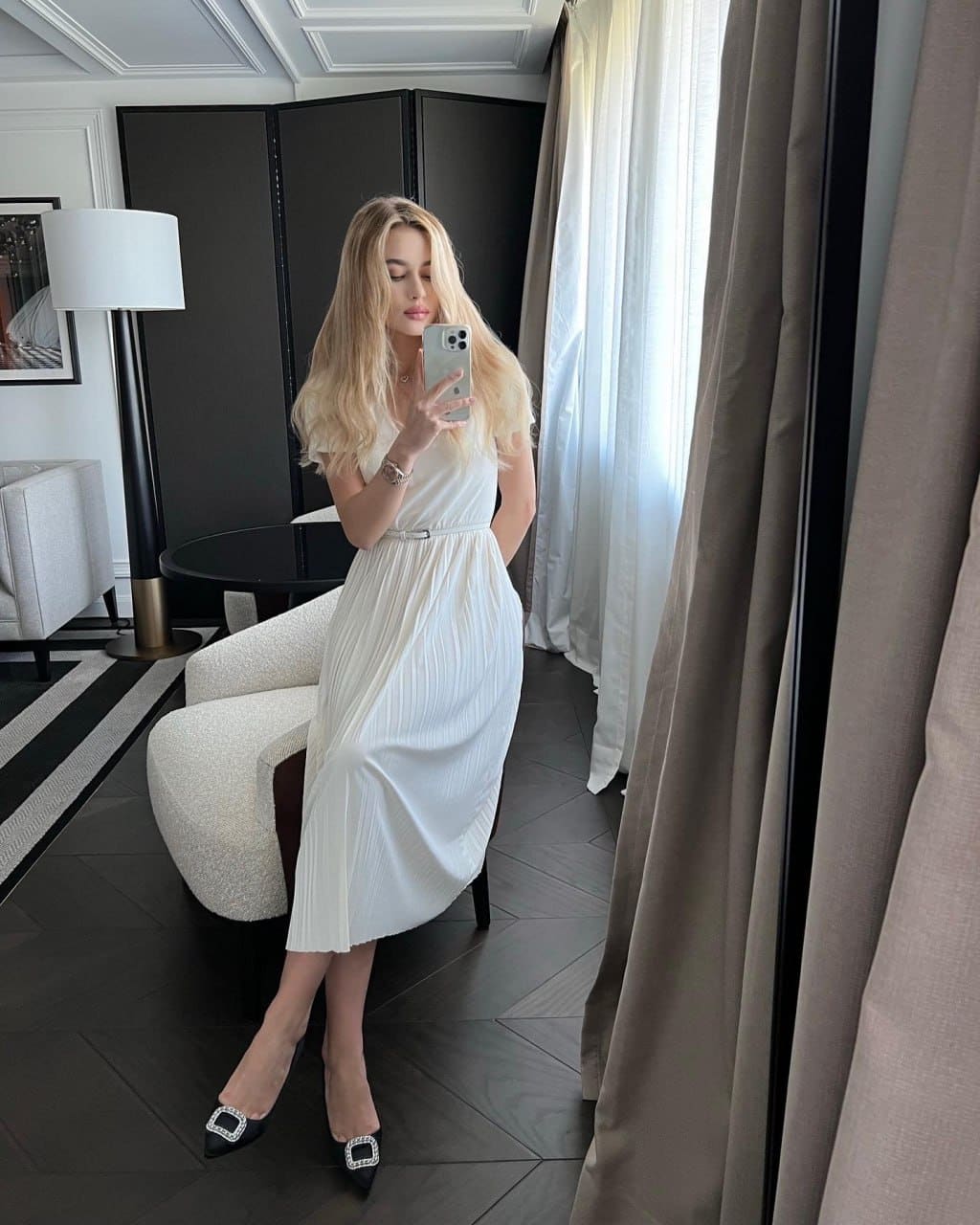A woman in a white pleated dress holds a phone in front of a mirror on dark wood floors.