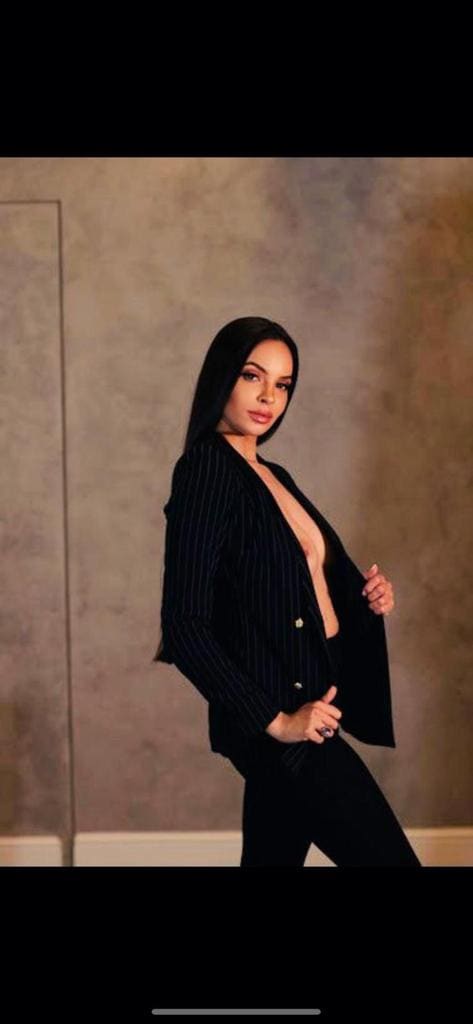 Mujer en pose de tres cuartos con blazer negro a rayas abierto y pantalones negros, ante pared texturizada.