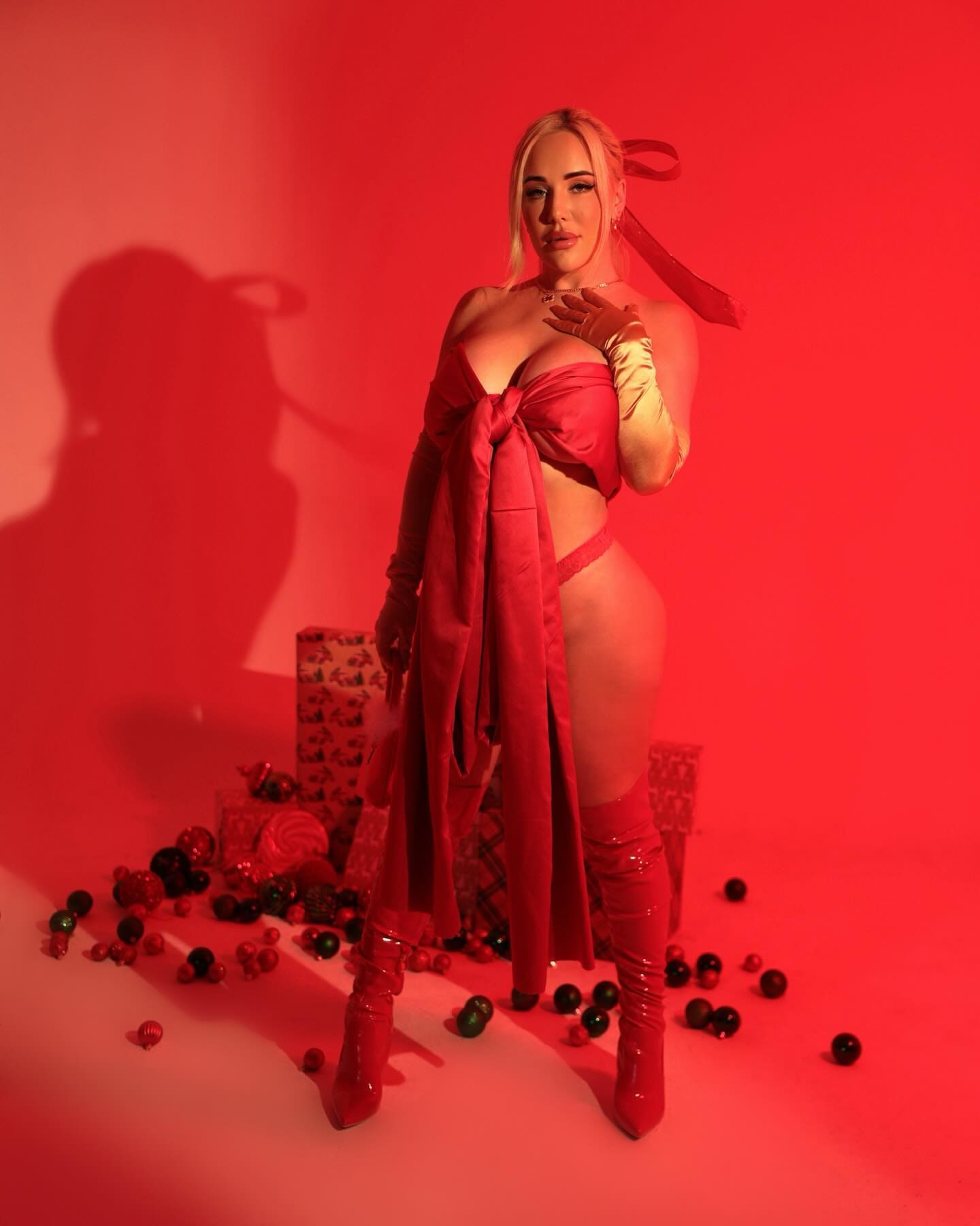 A blonde woman in red satin lingerie and gloves poses beside gifts in a red-lit studio setting.