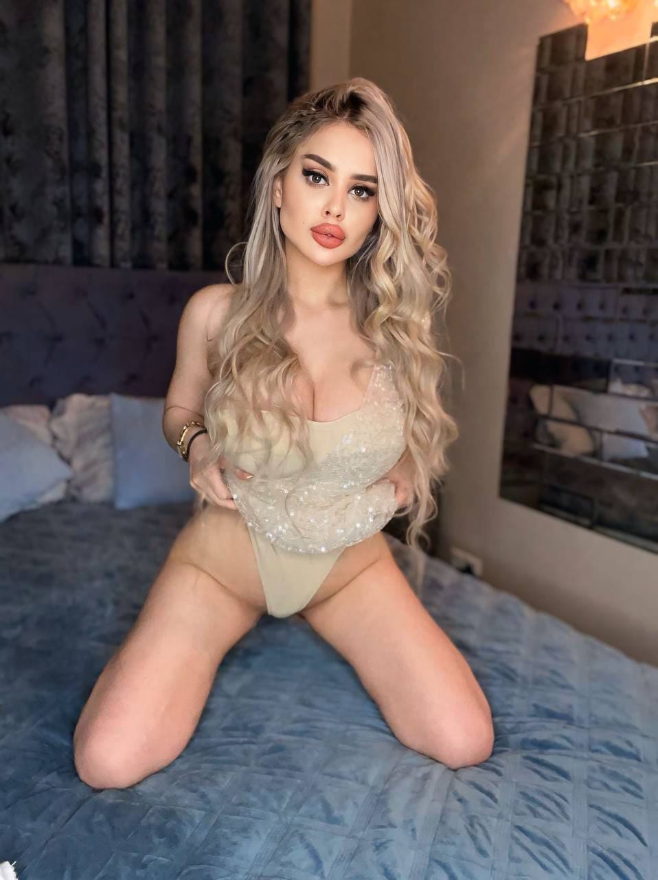 Mujer de pelo rubio ondulado arrodillada en cama azul con bodysuit beige de lentejuelas y escote profundo.