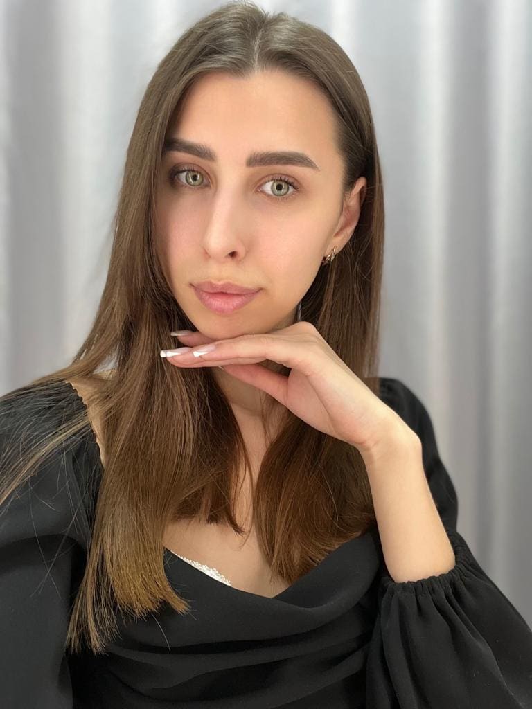 Karina con capelli castani lunghi appoggia il mento sulla mano, indossa un top nero a maniche a palloncino davanti a una tenda grigia.