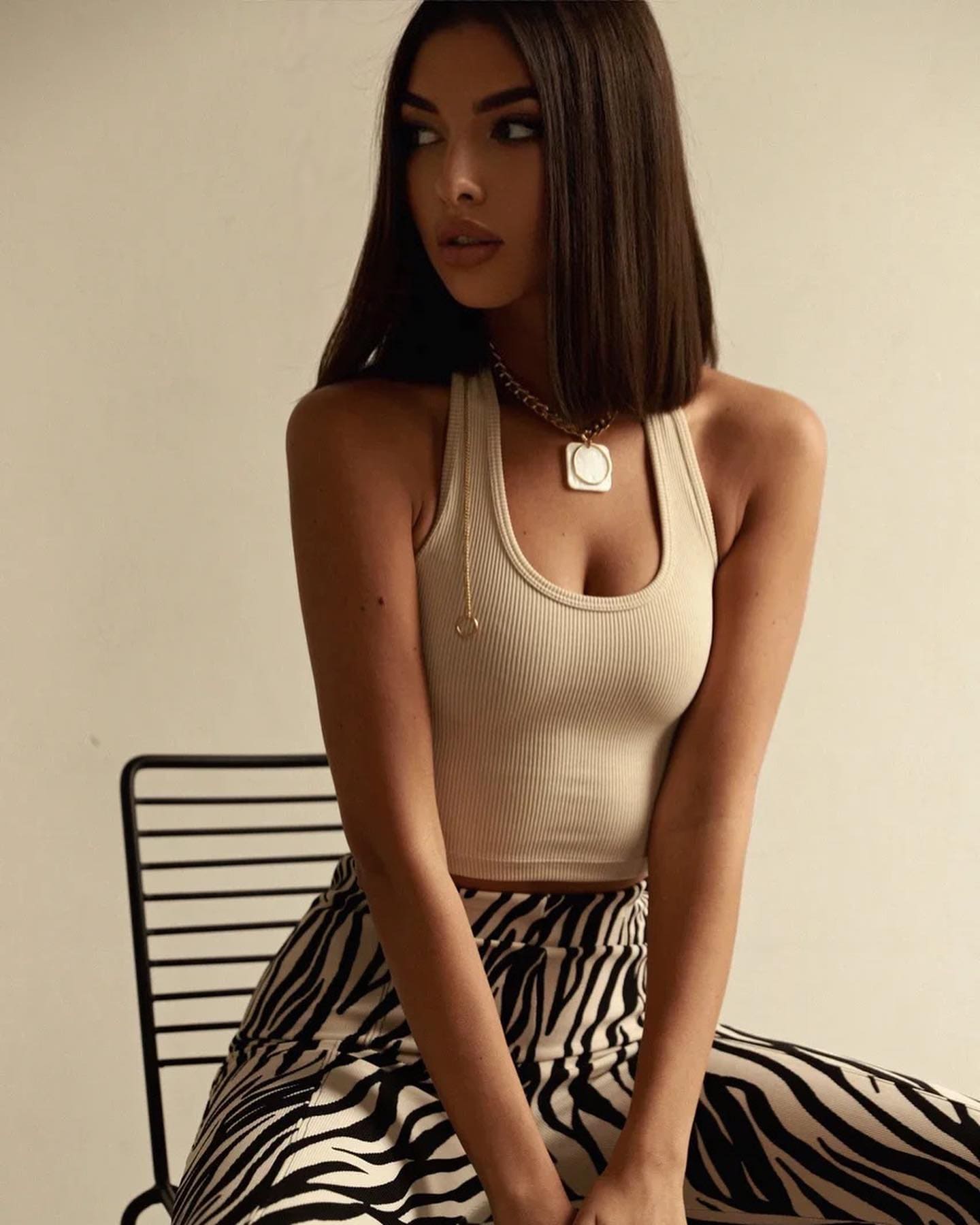 Mujer con cabello oscuro recto sentada en una silla metálica negra, usando top halter cremoso de punto y falda estampada cebra, con collar dorado contra una pared clara lisa.
