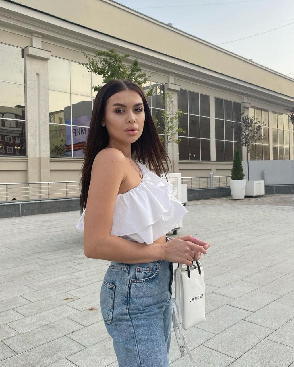 Mujer con cabello oscuro largo de pie en una plaza pavimentada frente a un edificio de cristal, vestida con top blanco rizado de un hombro y vaqueros azules claros, sujetando un bolso blanco pequeño.