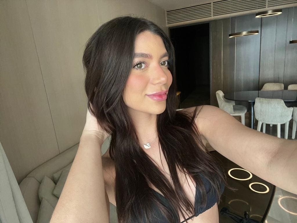 Selfie en interior moderno con top negro, mesa y sillas al fondo, brazo extendido.