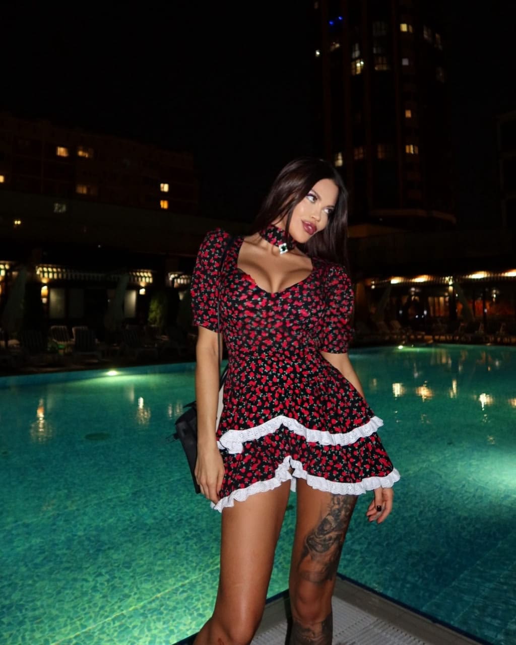 Donna con capelli scuri accanto a una piscina illuminata di notte, indossa un abito corto floreale rosso e nero con orlo bianco.