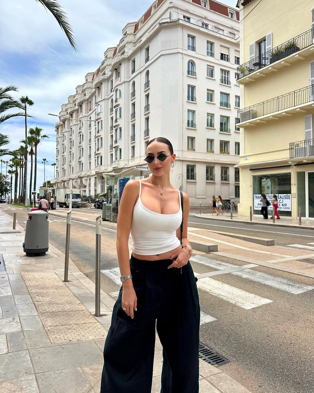 Mujer con top blanco, pantalón oscuro y gafas redondas en calle con edificios blancos.
