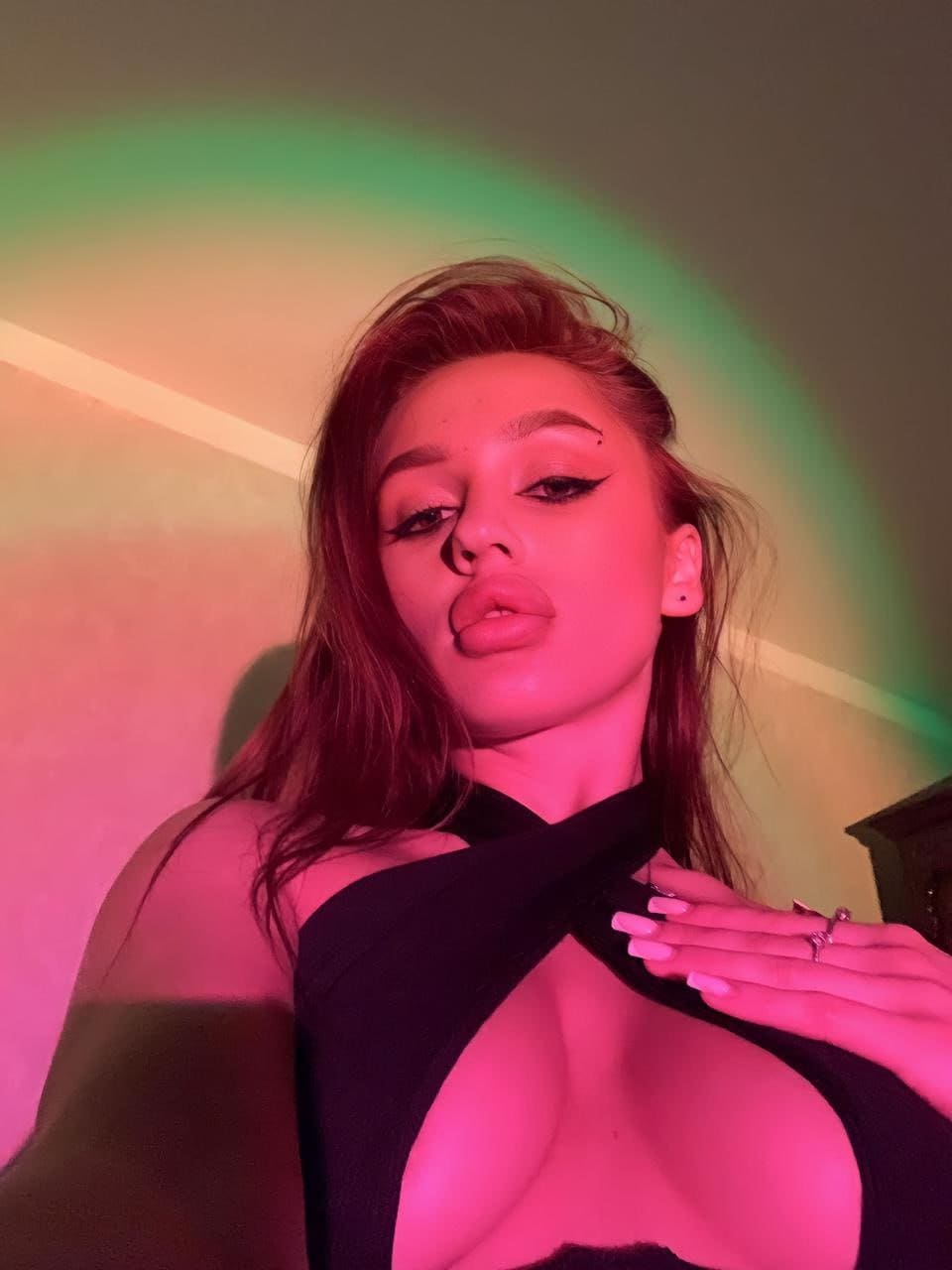 Mujer con cabello castaño rojizo y delineado alado posa con top negro halter bajo luces rosas y verdes frente a una pared.