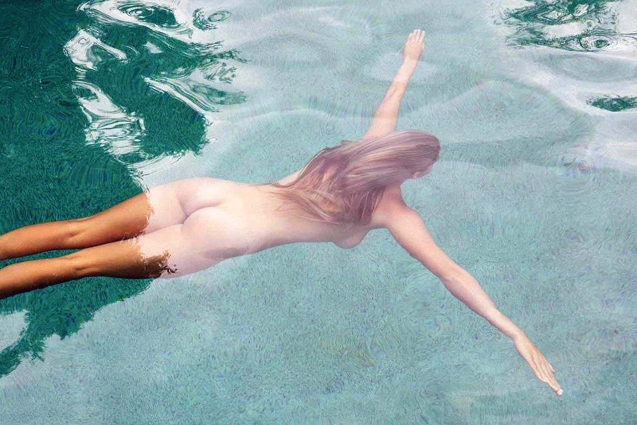 A person with long light hair floats face-down on a clear turquoise pool surface, arms extended forward.
