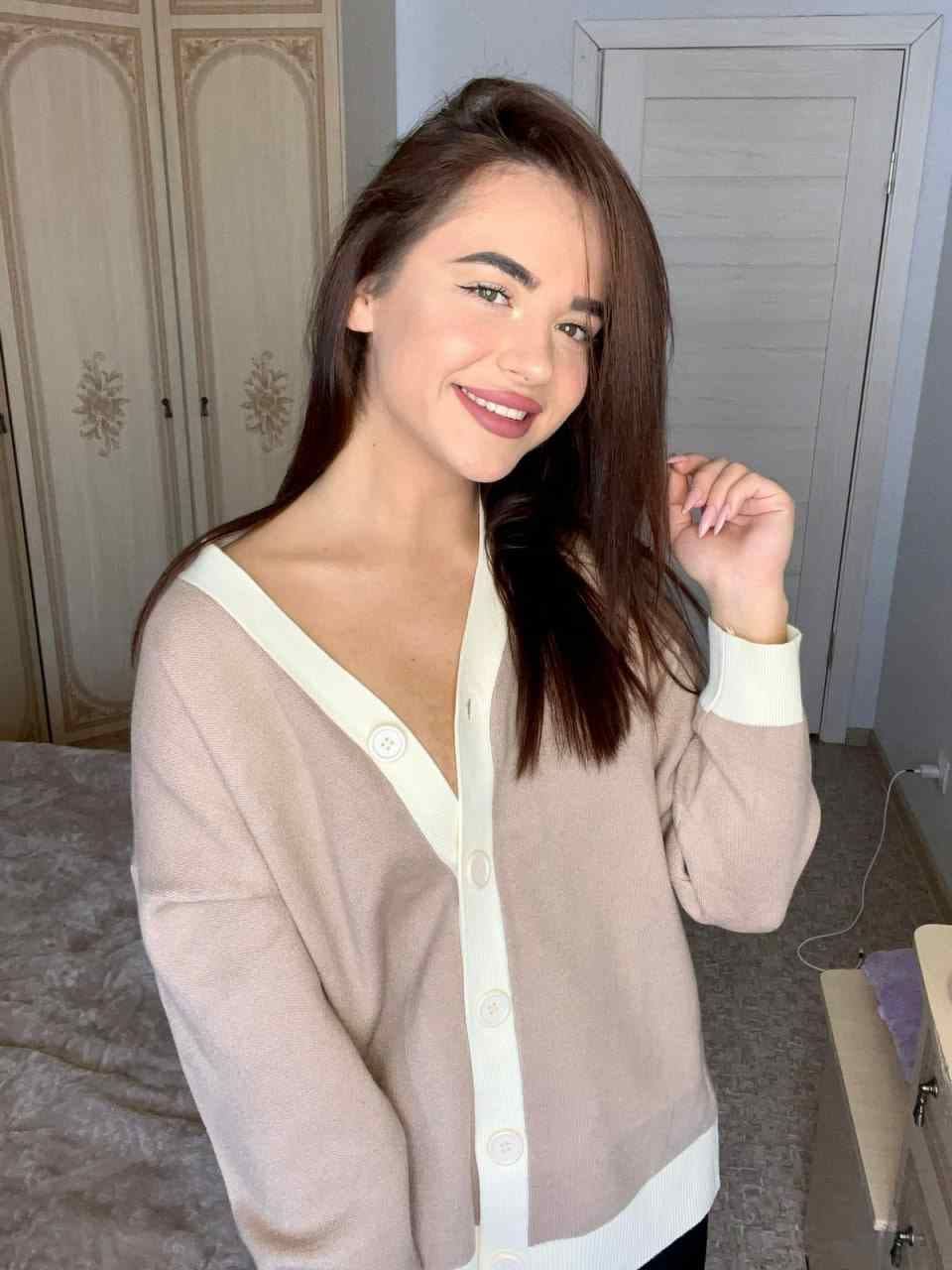 Karina con capelli scuri e lunghi, in un interno davanti a letto e armadio, indossa una cardigan beige chiaro con bordi bianchi.