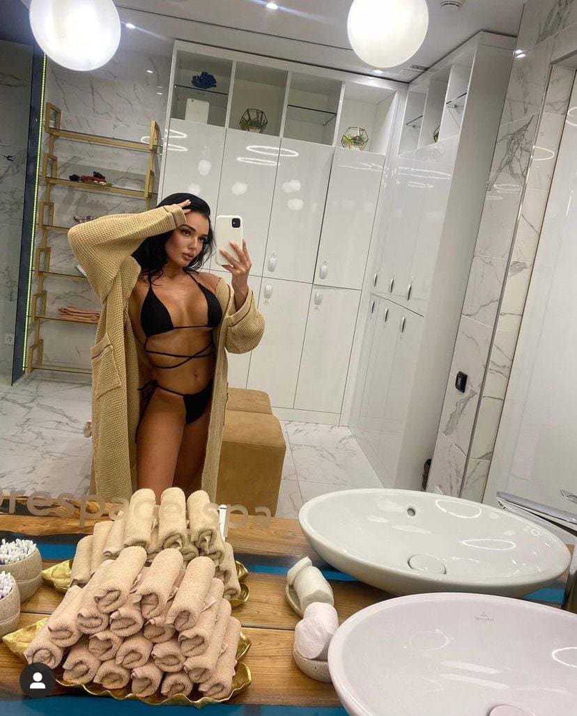 Mujer con bikini negra y cárdigan beige abierto haciendo selfie en el espejo de un baño de mármol con toallas y lavabos visibles.
