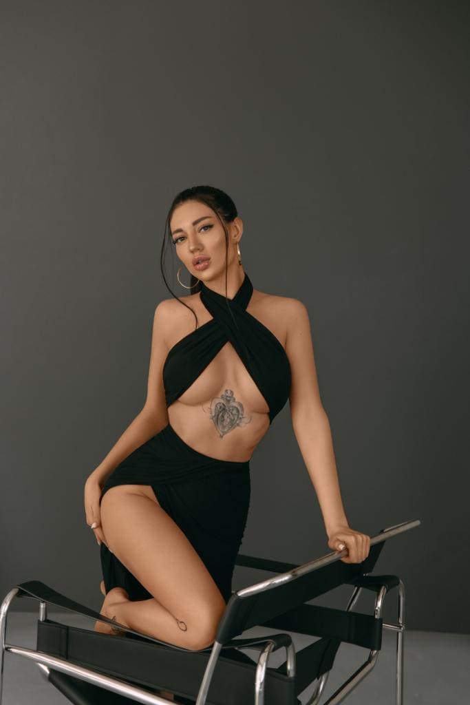 Mujer arrodillada sobre una silla negra y cromada, con top negro halter y falda corta a juego, ante fondo gris.