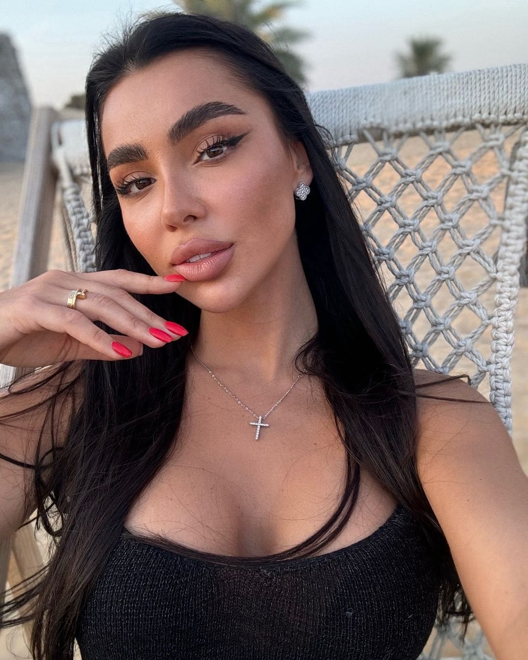 Marina con cabello oscuro y esmalte rojo posa en silla blanca tejida al aire libre, de top negro sin mangas, con palmeras al fondo.