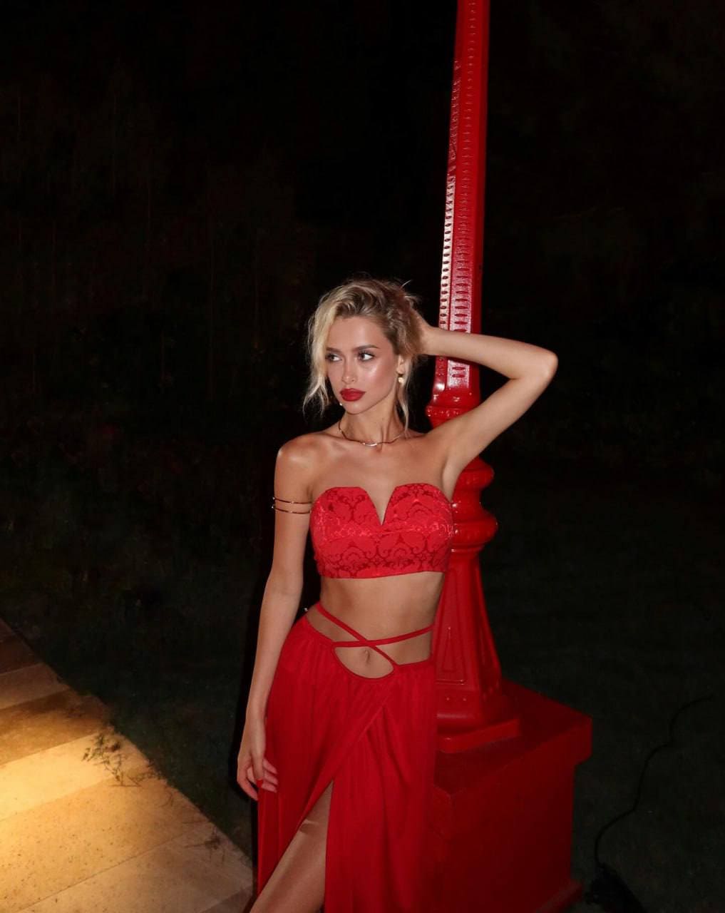 A woman in a red lace crop top and slit skirt stands beside a red pillar at night.