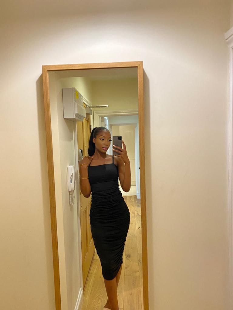 Donna con coda di cavallo in abito nero senza maniche che si fa uno specchio selfie in un corridoio con pavimento in legno.