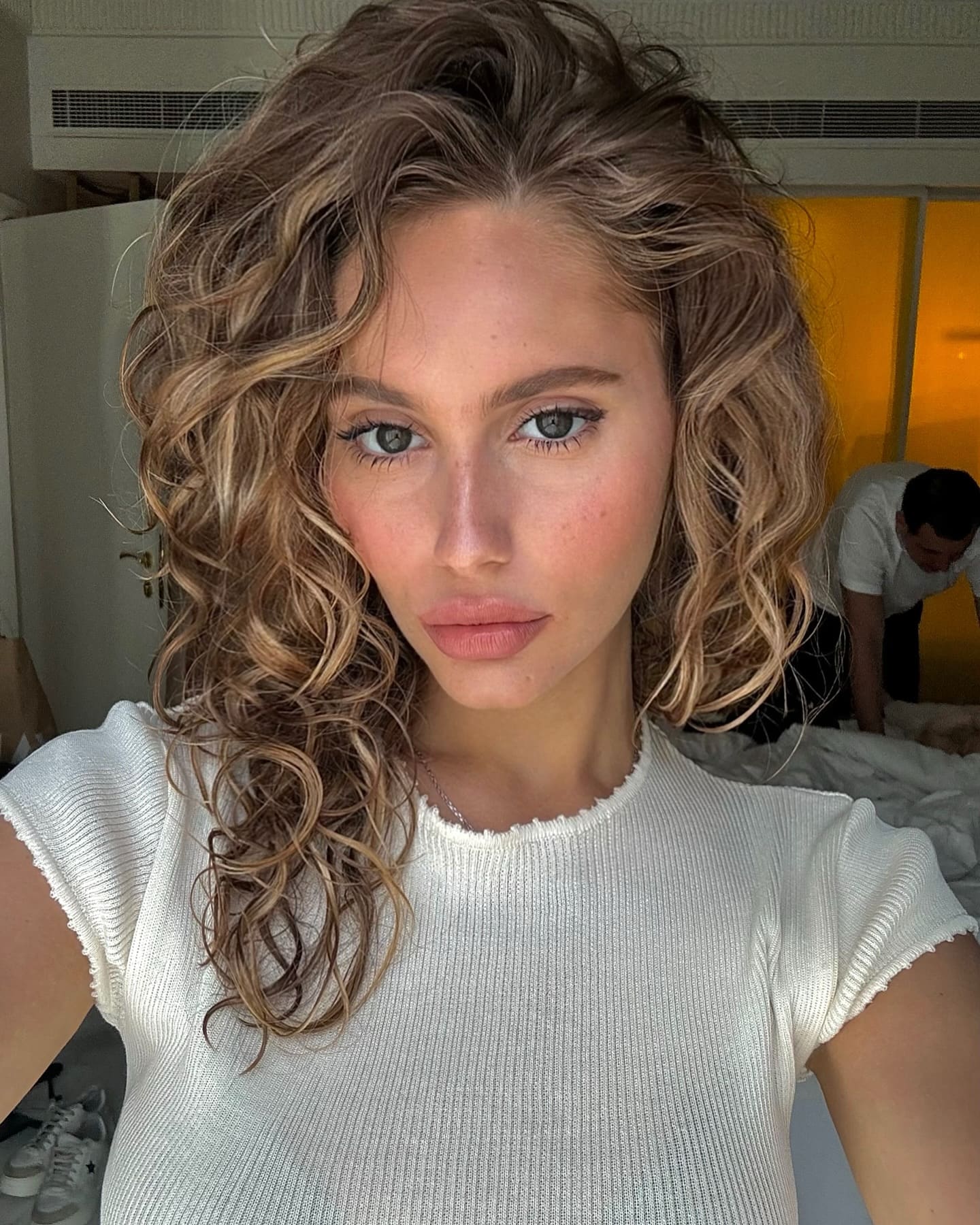 ميمي بشعر أشقر طويل مموج تلتقط صورة سيلفي بقميص أبيض قصير الأكمام في غرفة يوجد فيها سرير.