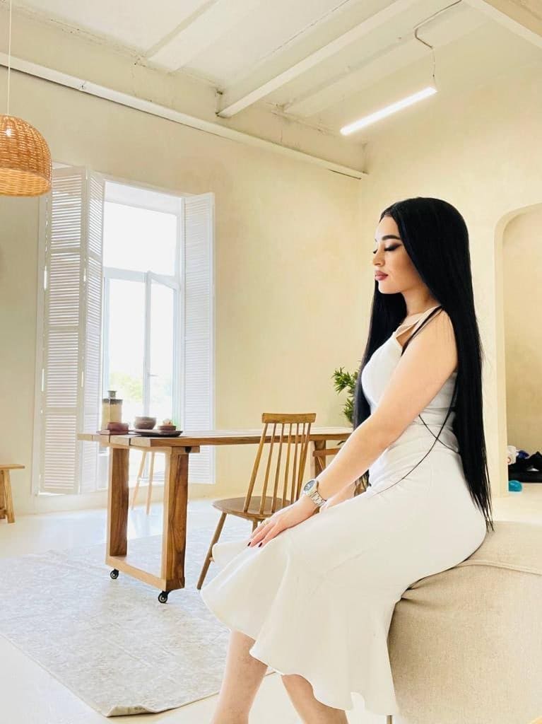 Samira sits in profile on a beige sofa in a white midi dress, in a bright room with wooden furniture and white shutters.
