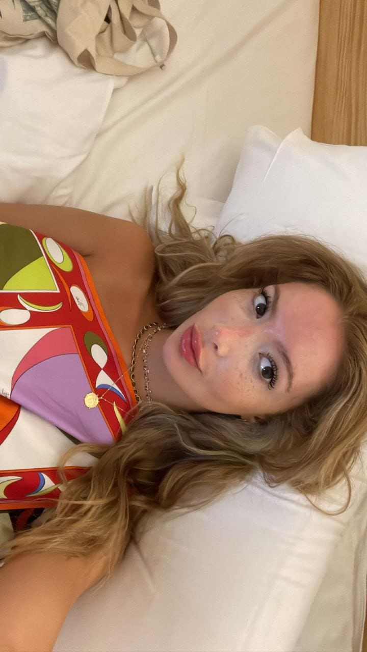 Clara lying on a white bed in a patterned strapless top and gold necklace.