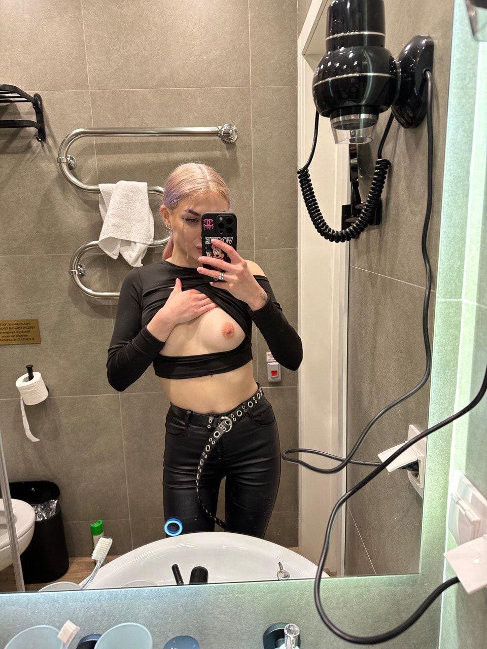 Rubia en baño de azulejos subiendo el top negro de mangas largas para exponer un pecho, con pantalones de cuero negros.
