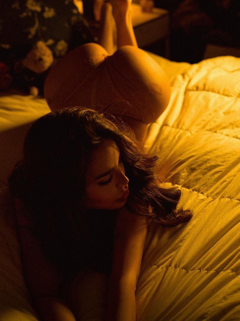 Mujer tumbada de lado en cama con edredón amarillo, vestido claro corto y piernas flexionadas hacia atrás en habitación oscura.