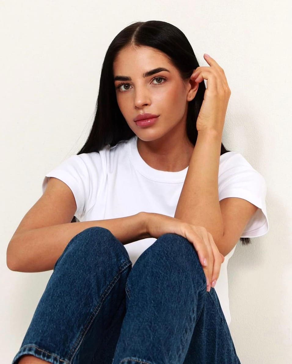 Anastasia sits with knees drawn up in a white t-shirt and jeans, one hand raised to her hair against a light wall.