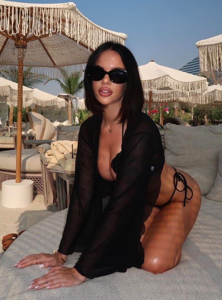 Mujer arrodillada en cojín gris con gafas, bikini negro y camisa transparente larga bajo palmeras y sombrillas.
