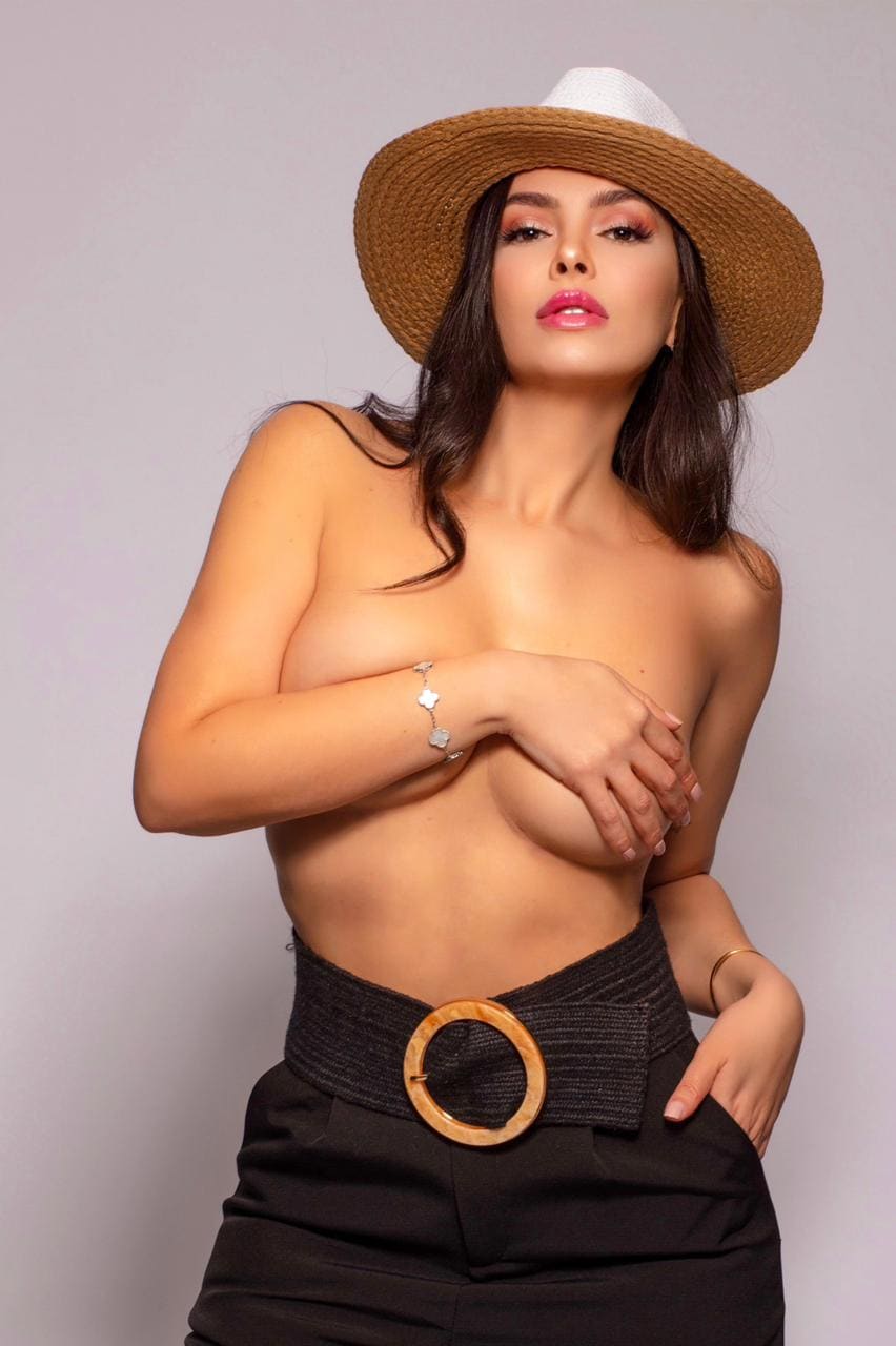 A woman wears a straw hat and high-waisted trousers, posing topless with one arm crossed against a gray wall.