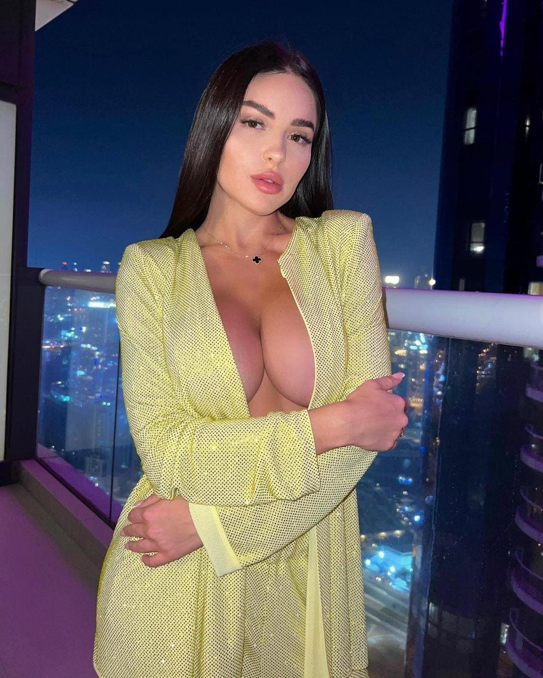Mujer con cabello oscuro en balcón nocturno, vestida con vestido amarillo perforado y cuello V profundo, brazos cruzados.