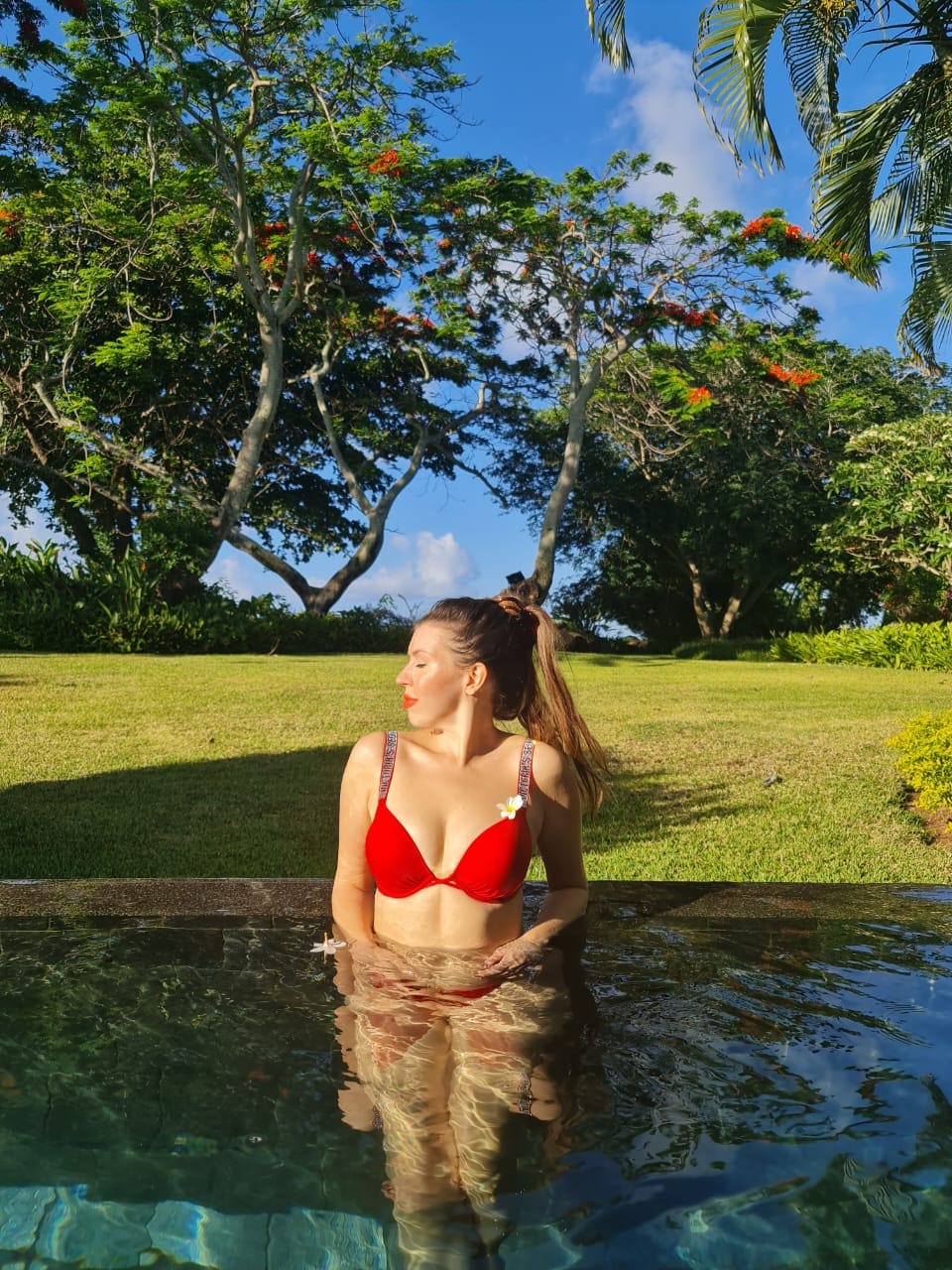 Mujer con coleta alta en una piscina, vistiendo top de bikini rojo y flor blanca detrás de la oreja, bajo cielo azul.