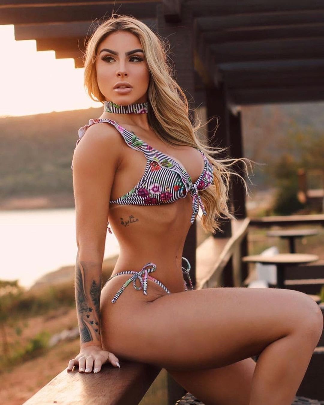 Rubia sentada en banco exterior con bikini floral y gargantilla, brazo izquierdo apoyado y tatuajes visibles en antebrazo derecho.