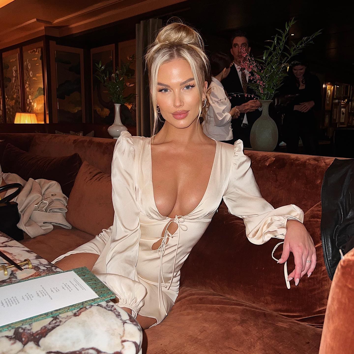 Rubia con moño alto y vestido crema de manga larga con escote V se sienta en un sofá de terciopelo marrón en una sala tenue.