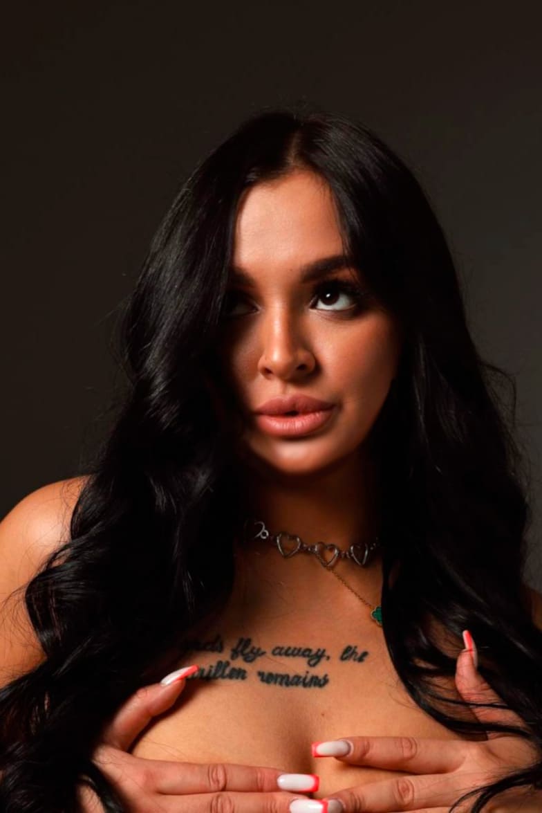 Mujer con hombros descubiertos y collar de corazones, tatuaje en pecho superior y uñas rojas, manos sobre el busto.