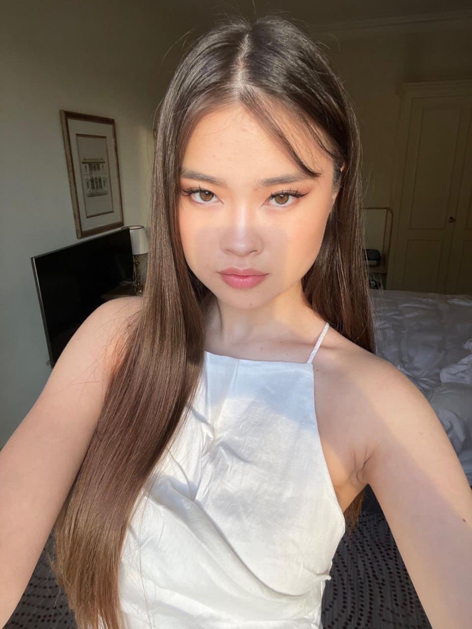 A young woman takes a selfie in a bedroom wearing a white halter top, with a bed and framed picture behind her.