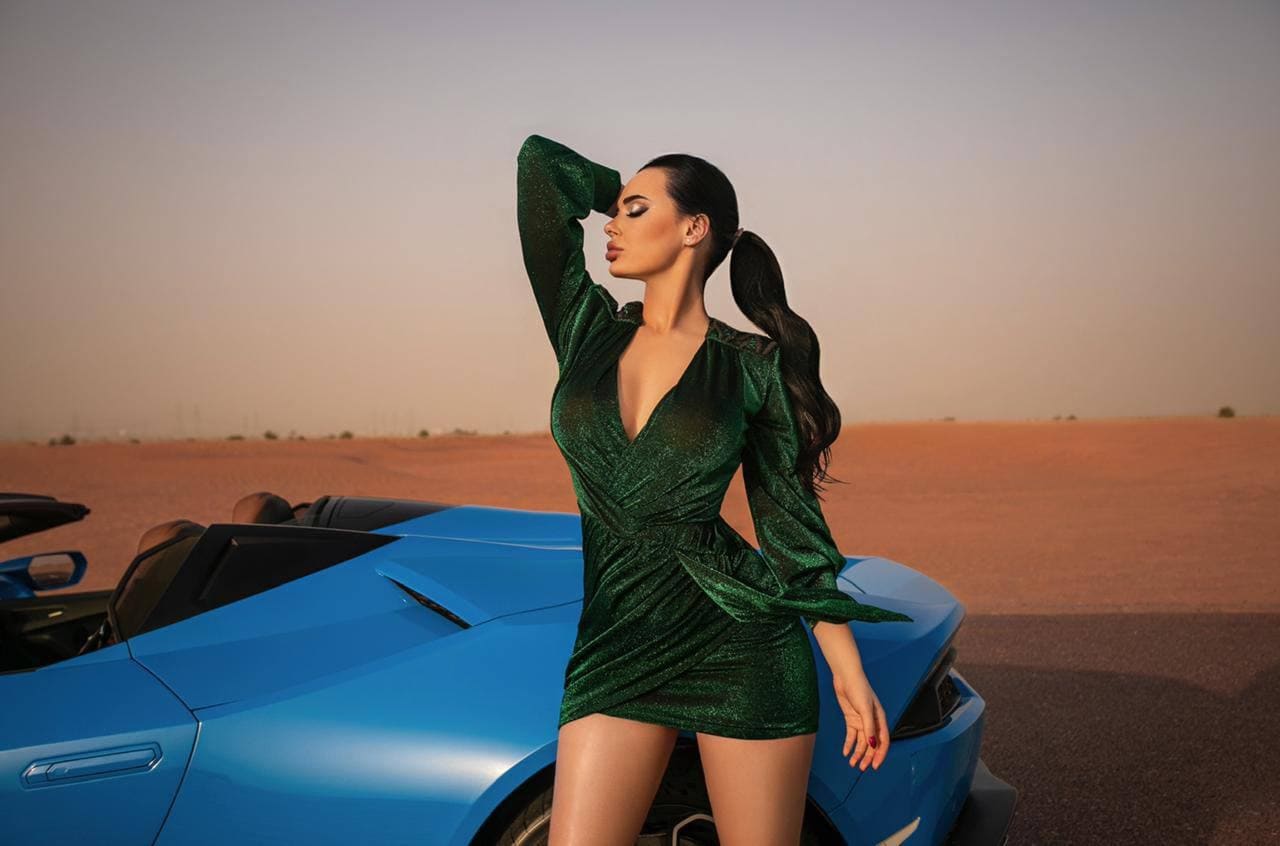 Mujer de cabello oscuro junto a un descapotable azul en el desierto, vestida de verde y mano en la frente.