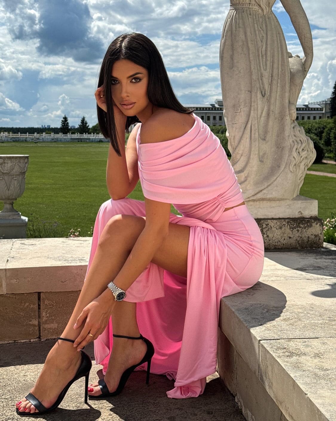 Mujer con cabello largo oscuro sentada en un borde de piedra, vistiendo un vestido rosa descubierto en los hombros y tacones negros, con una estatua y césped cuidado al fondo.