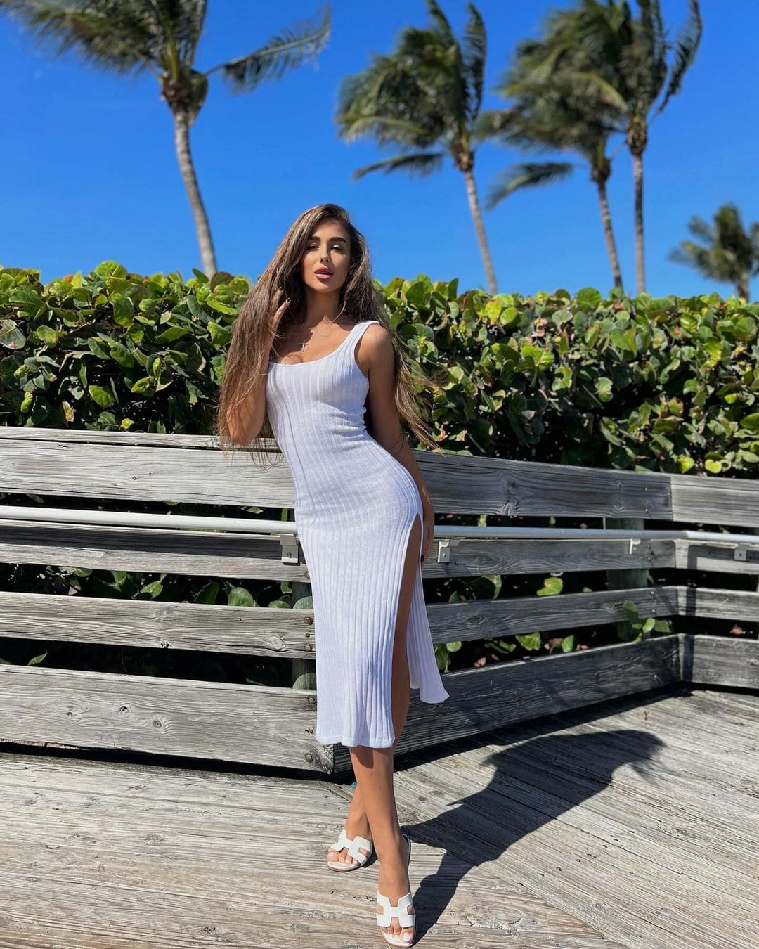 A woman in a white ribbed midi dress leans against a wooden railing on a deck with green hedges and palms behind.