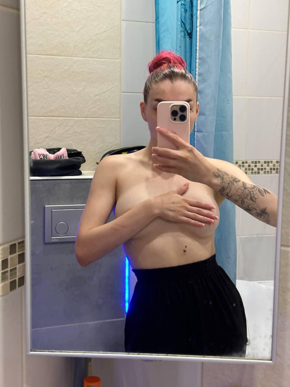 Persona con moño rosa en baño cubriendo el pecho con una mano y sosteniendo el teléfono, vistiendo falda negra.