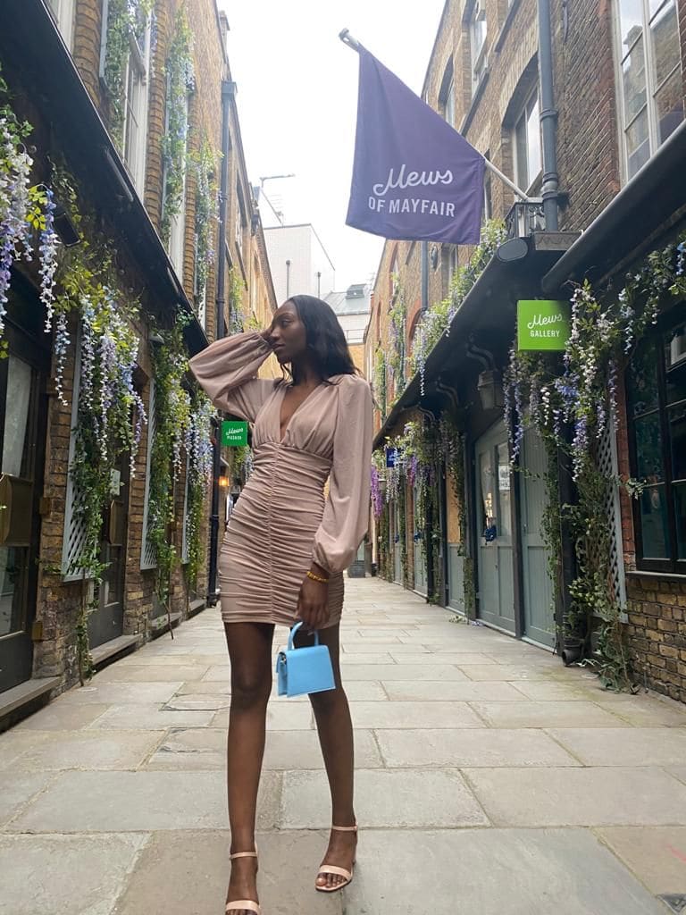 Donna in vicolo di mattoni con glicini, abito beige attillato e sandali alti, tiene una borsa azzurra in mano.