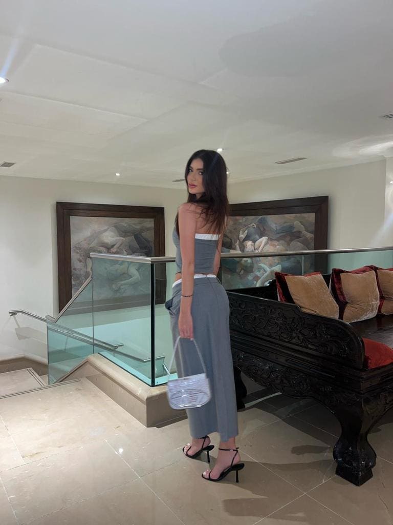 En vestíbulo con conjunto gris, bolso transparente, tacones negros y barandilla.