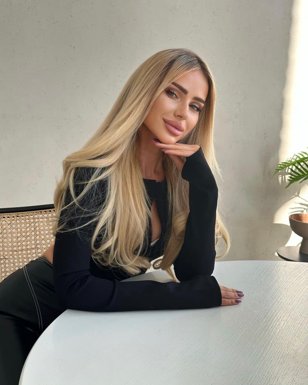 A woman sits at a table in a black long-sleeved top, resting her chin on one hand with a plant in the background.